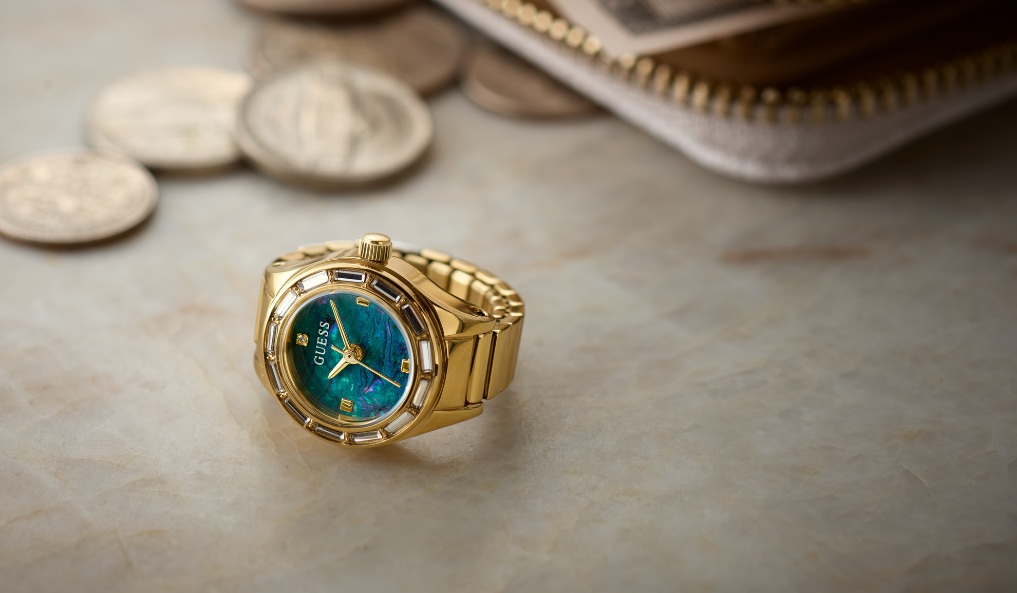 A gold-tone GUESS watch ring with a blue dial and bezel, styled on a light marble surface with coins and a wallet in the background.