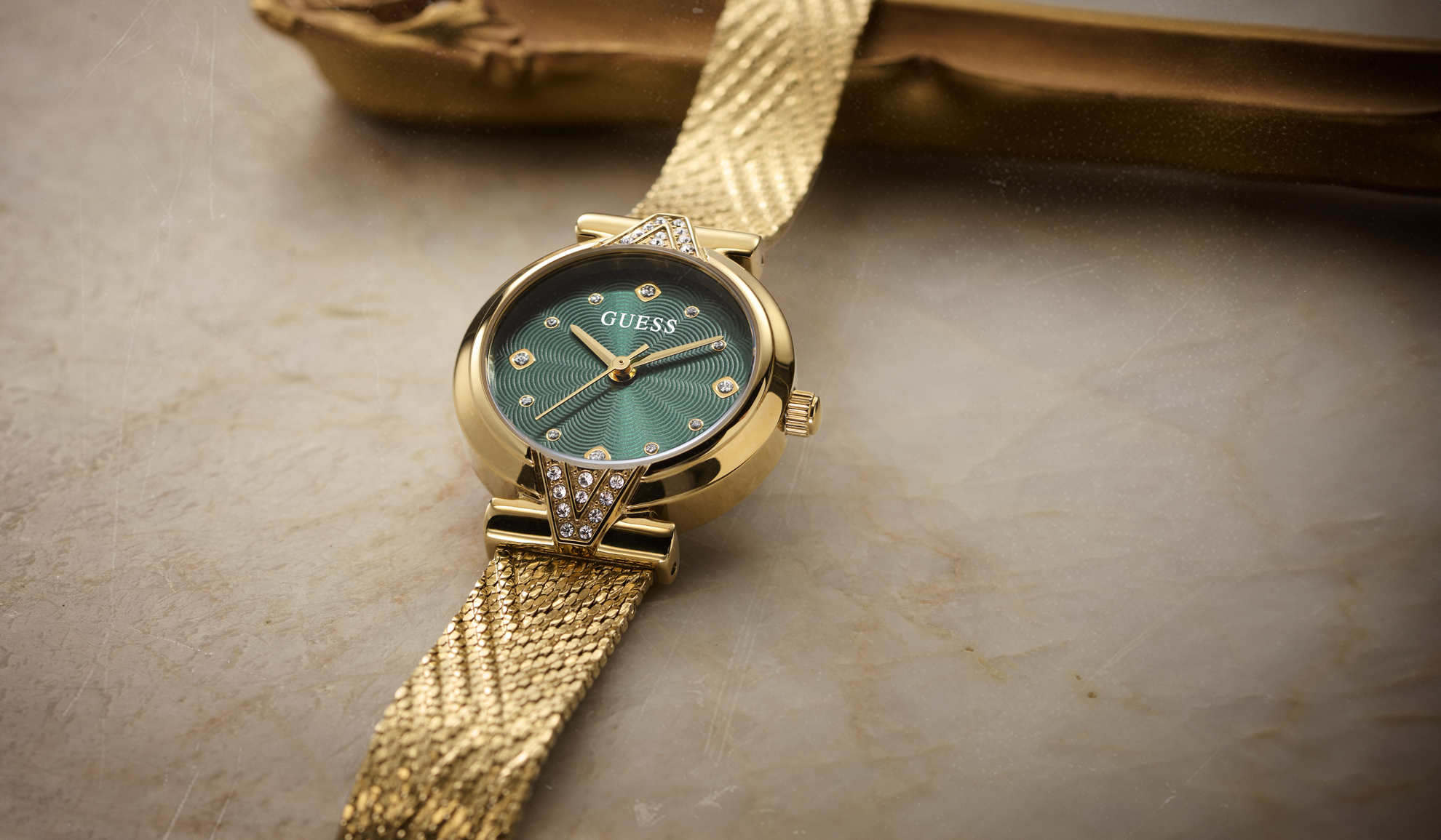 A gold-tone Guess wristwatch with a textured mesh strap, a green dial, and crystal hour markers, resting on a beige stone surface.