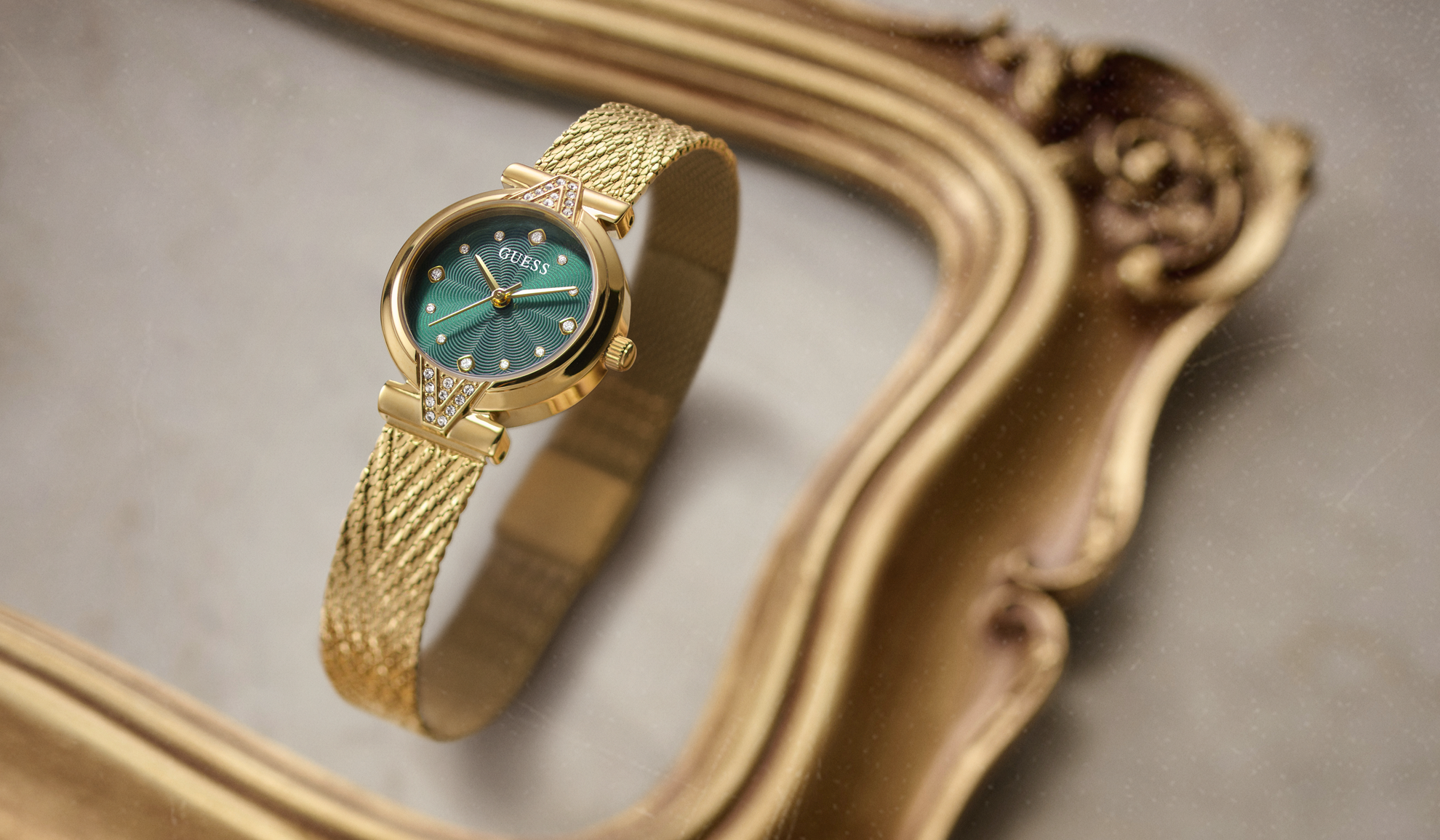 A GUESS wristwatch with a gold mesh band and a green dial, resting on an ornate gold mirror frame against a neutral gray background.