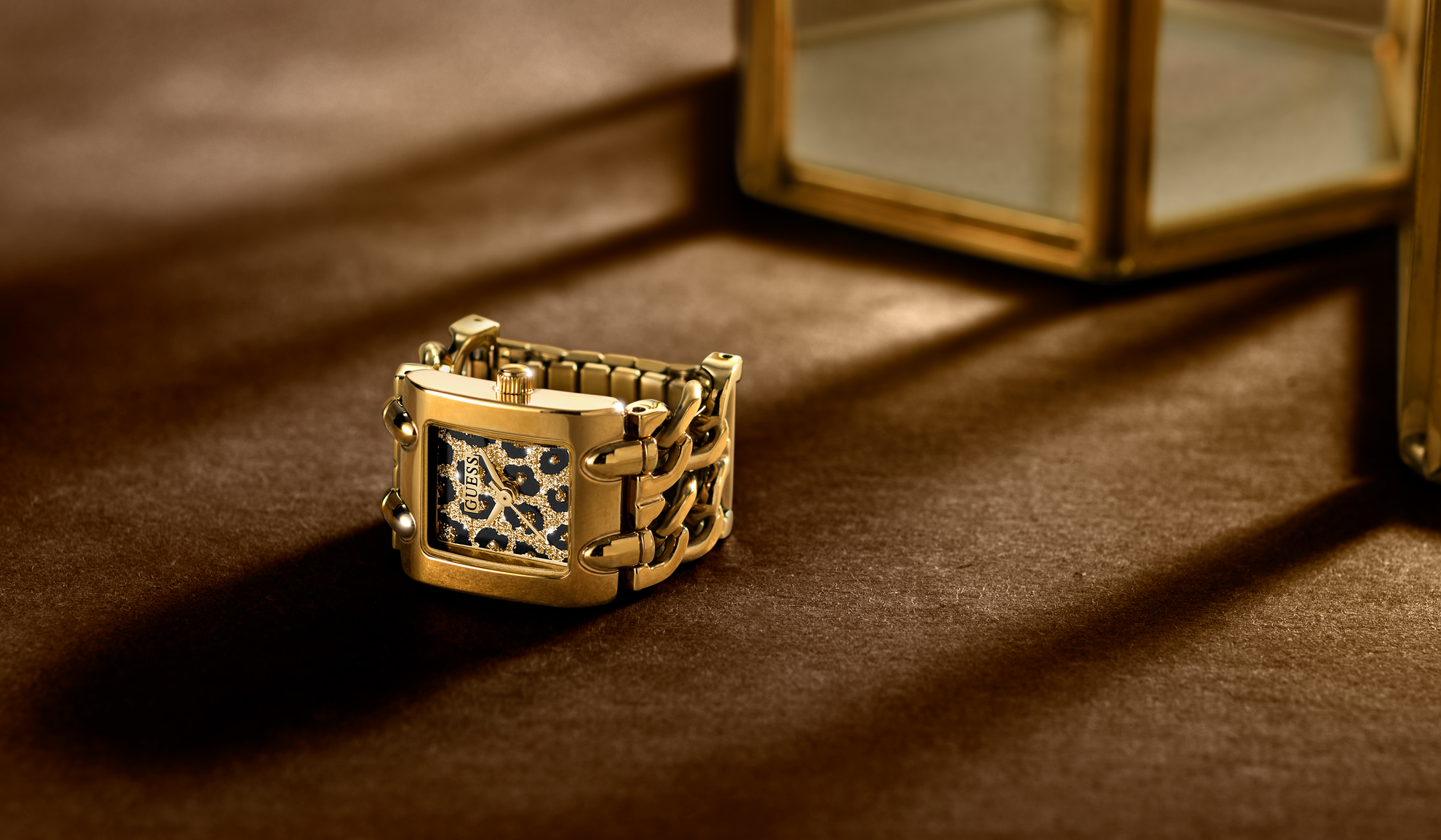A gold-tone watch ring with a rectangular case and a patterned dial, resting on a dark brown textured surface with dramatic shadows.