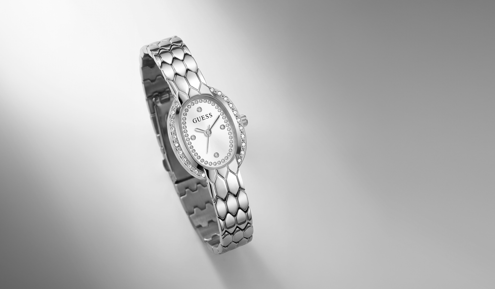 A silver-tone GUESS watch with a textured link bracelet and a diamond-encrusted bezel, shown floating against a clean gray gradient background.