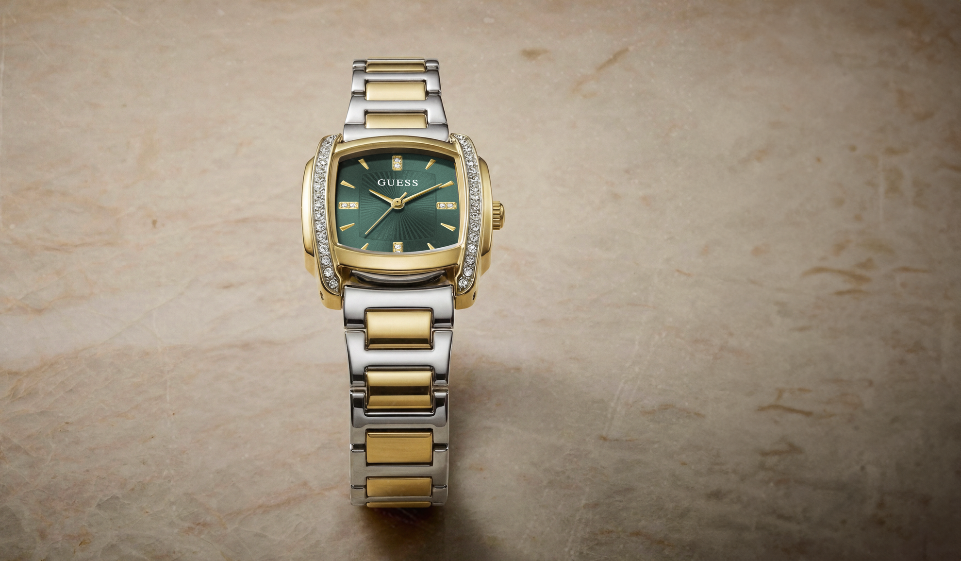 A GUESS two-tone watch with a green dial, gold-tone case accents, and a silver-and-gold link bracelet, displayed on a textured beige stone surface.