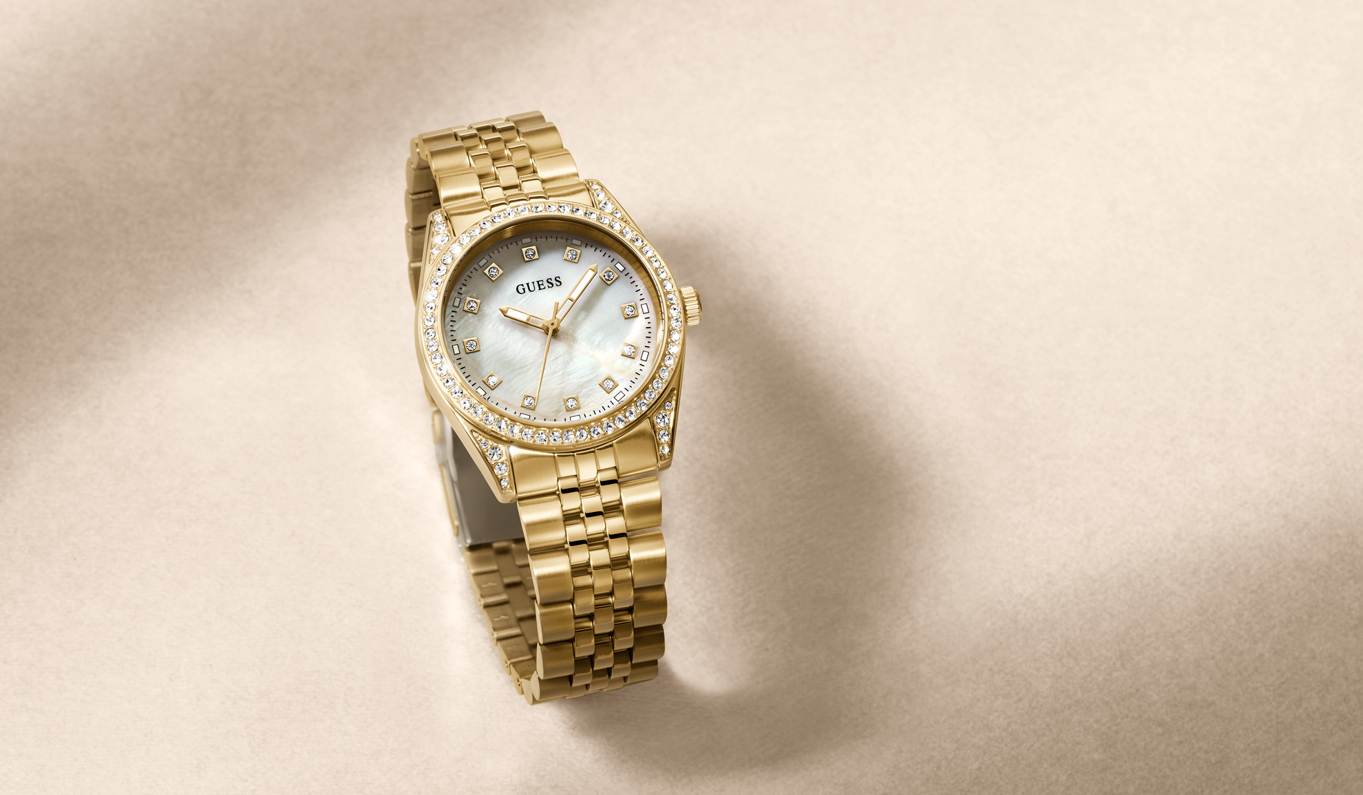 A gold-tone Guess wristwatch with a diamond bezel and a mother-of-pearl dial, displayed on a textured beige background.