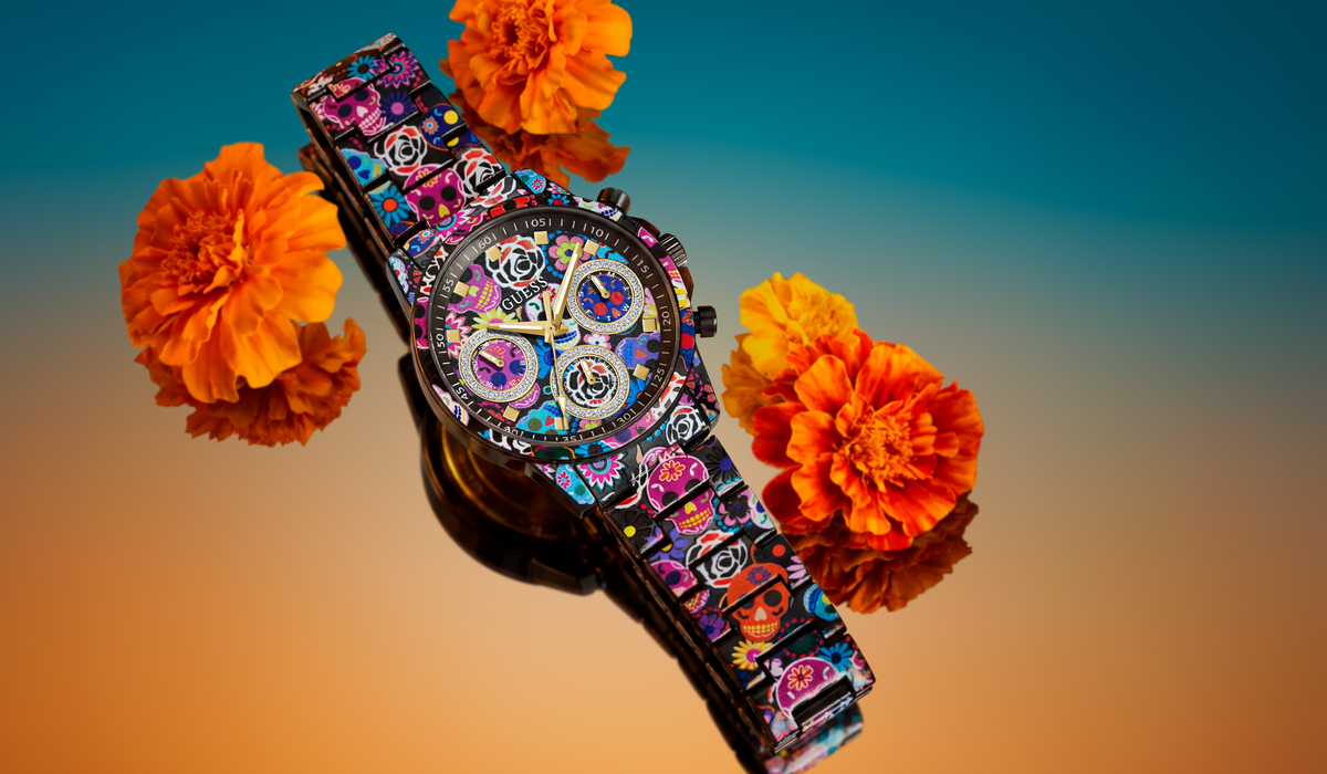A colorful wristwatch with a patterned strap and a detailed multicolor dial, styled with orange marigold flowers against a blue-to-orange gradient background.