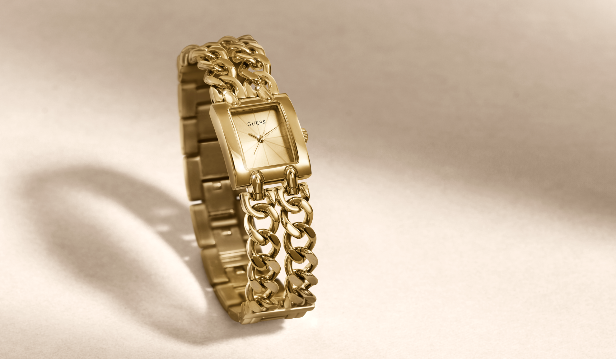 A gold-tone GUESS watch with a rectangular dial and a chunky chain-link bracelet, photographed on a warm beige background with a soft shadow.