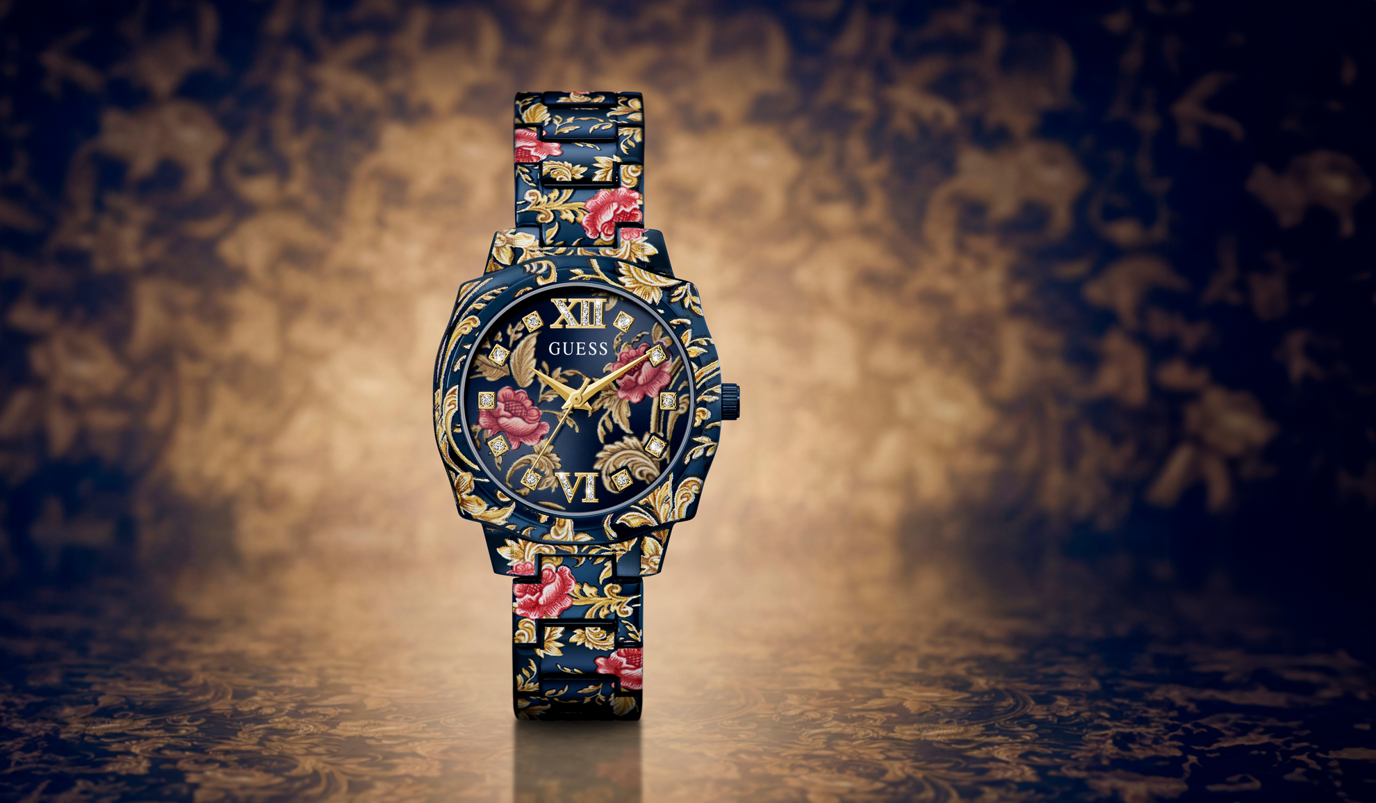A GUESS wristwatch with a dark blue floral patterned case and strap, featuring gold-tone hands and Roman numeral hour markers, set against a dark background with a warm spotlight effect.