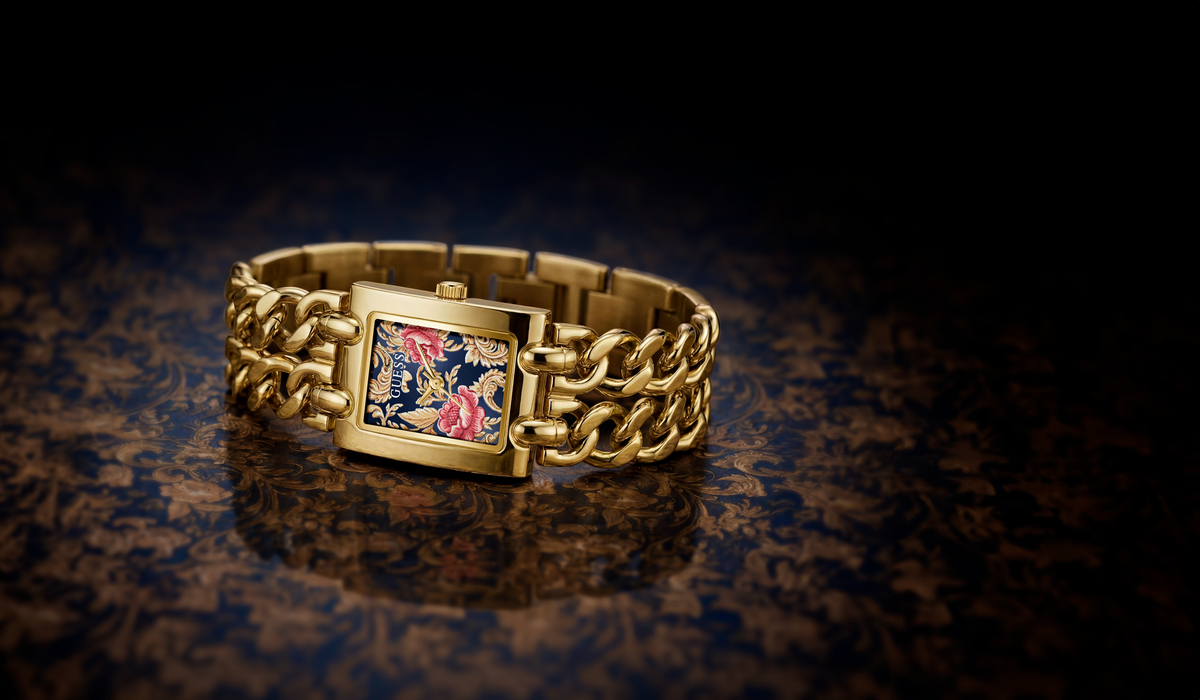 A gold-tone metal bracelet watch with a rectangular face featuring a colorful floral pattern, resting on a dark reflective surface.