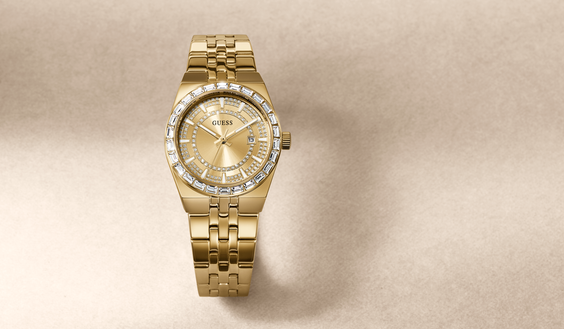 A gold-tone Guess watch with a diamond-encrusted bezel and a gold dial, shown on a textured beige background.