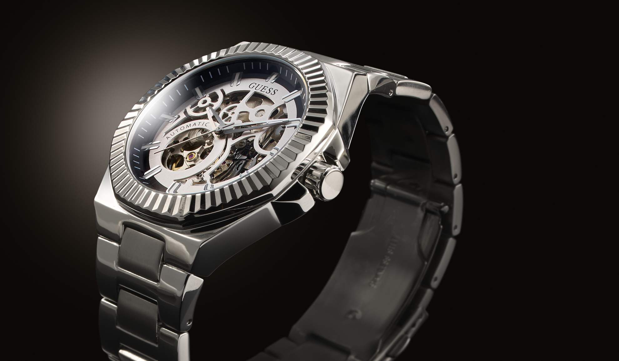 A close-up studio shot of a silver-tone skeleton wristwatch with a matching metal link bracelet, set against a dark black background.