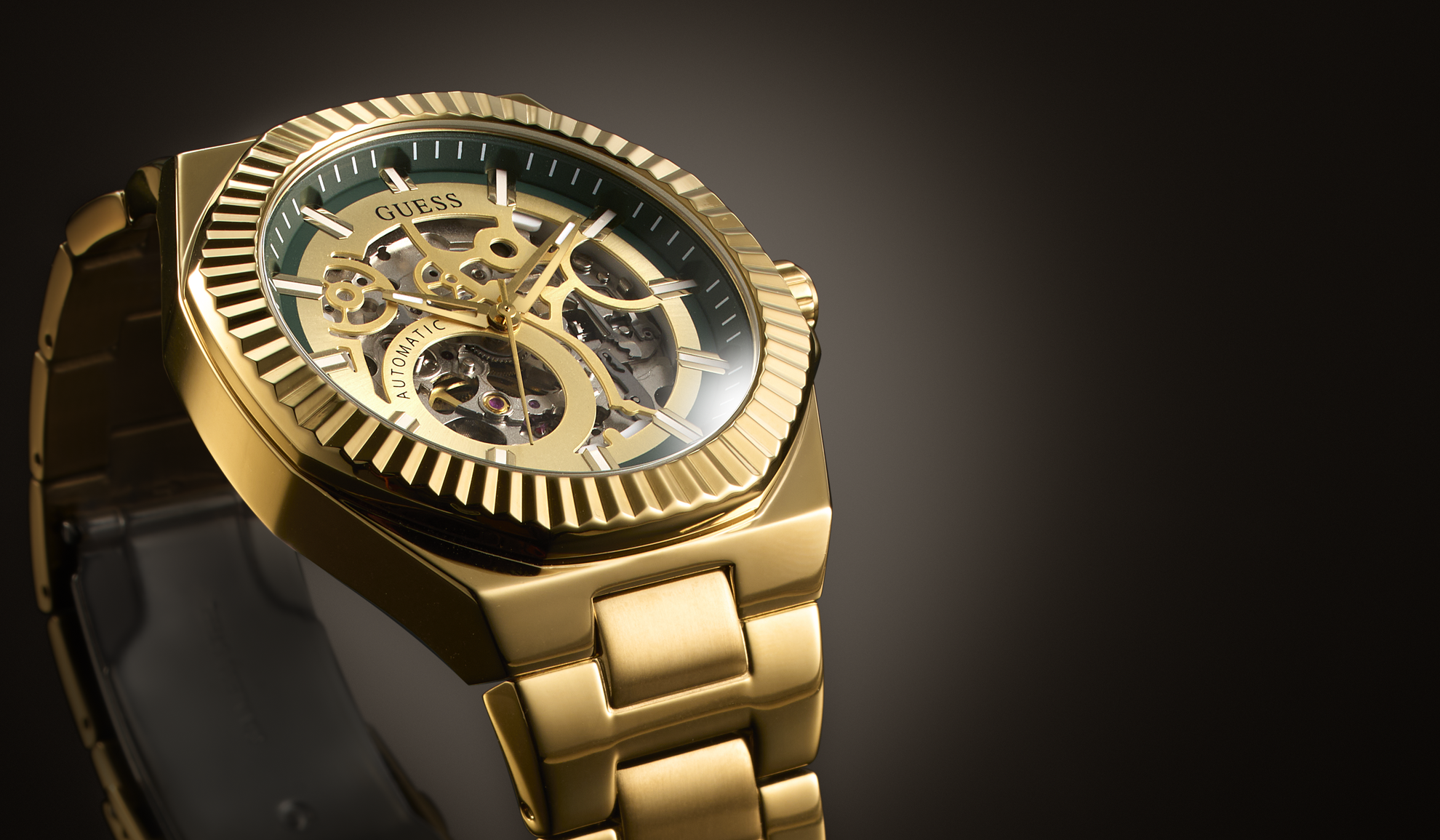 Gold-tone Guess wristwatch with a skeleton dial, fluted bezel, and matching metal link bracelet, shown on a dark gradient background.
