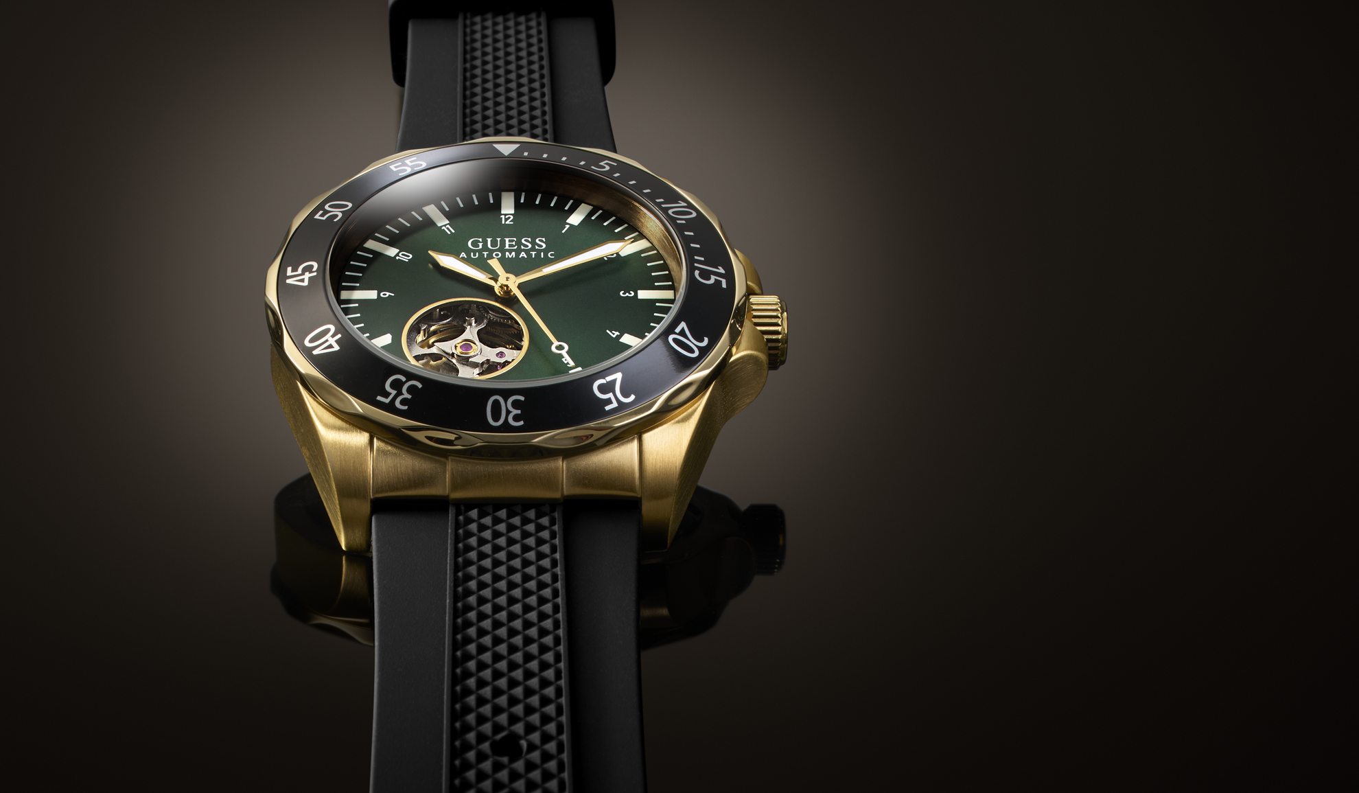 A Guess wristwatch with a gold-tone case, dark green dial, black bezel, and black textured strap, shown against a dark gradient background.