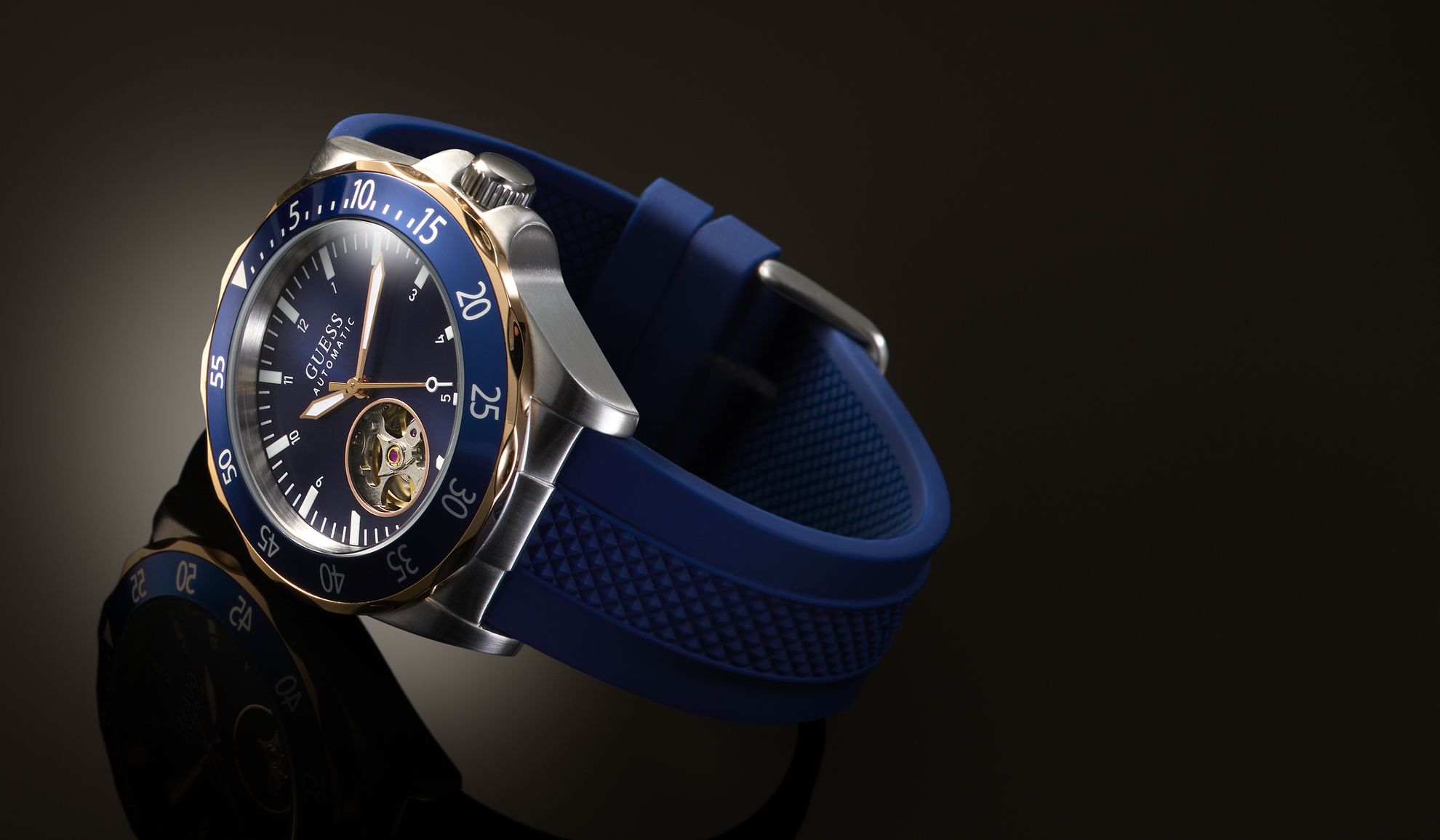 A luxury wristwatch with a blue textured silicone strap, a blue dial, a gold-tone bezel, and a silver-tone case, shown on a dark reflective surface.