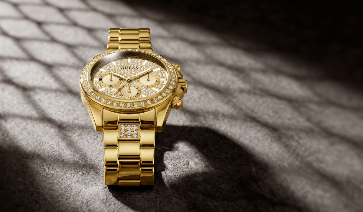 A gold-tone GUESS chronograph watch with a diamond-encrusted bezel and matching diamond-set bracelet, displayed on a textured gray surface with dramatic diagonal shadows.