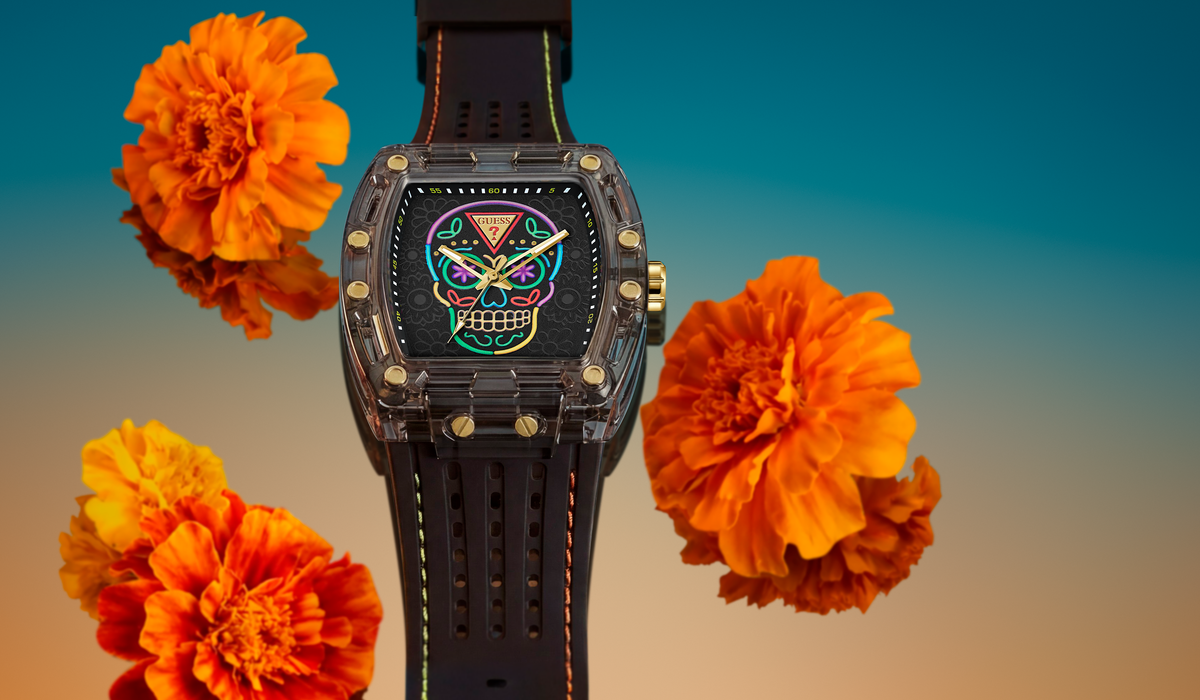 A GUESS watch with a black strap and a clear case, featuring a colorful sugar skull dial design. The watch is centered against a gradient background of blue and orange, surrounded by three orange marigold flowers.