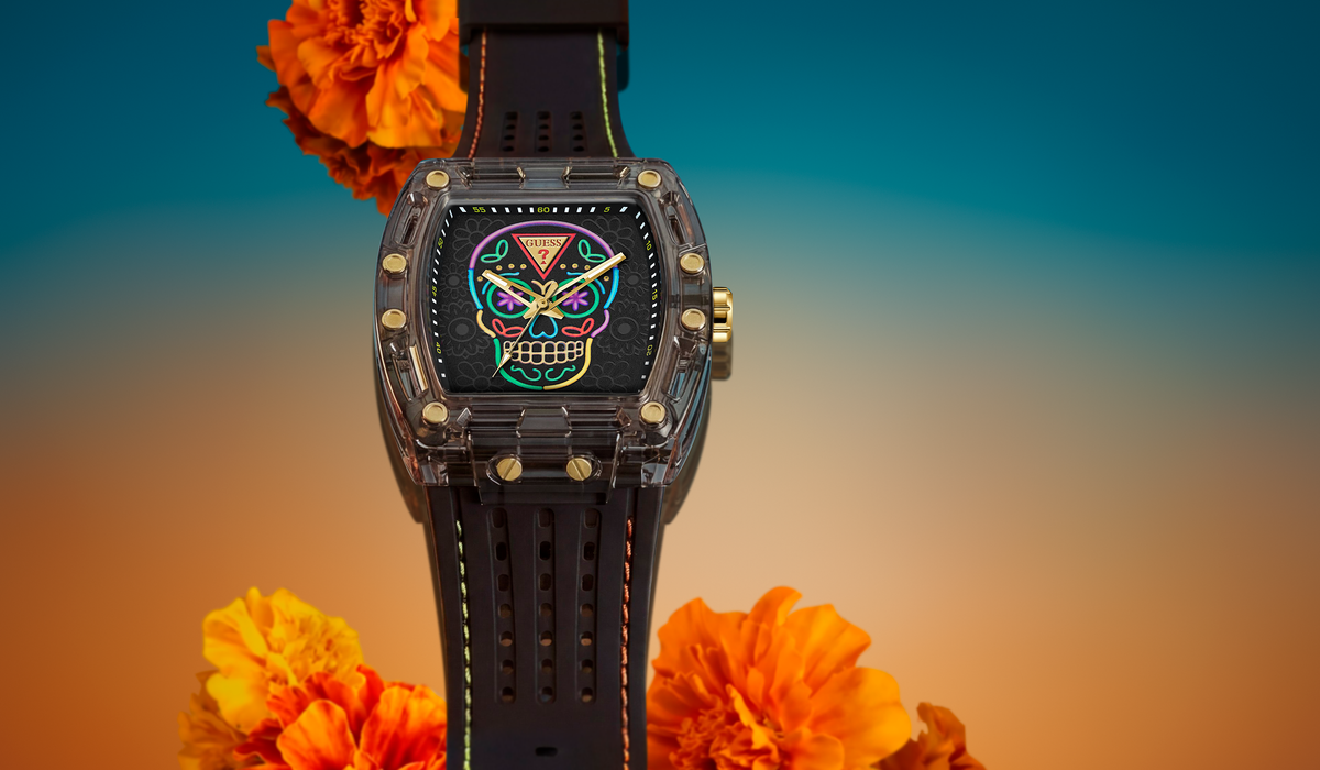 A luxury watch with a black strap and a transparent case, featuring a colorful sugar skull dial design. The watch is styled with orange marigold flowers against a blue-to-orange gradient background.