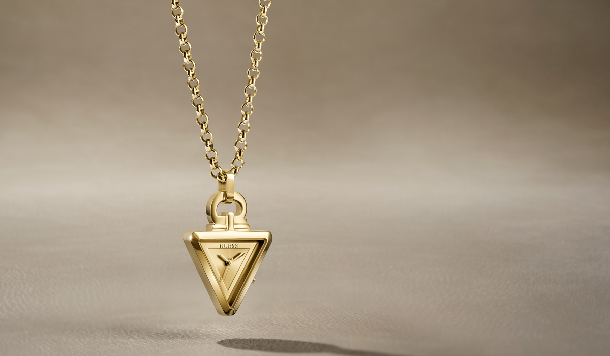 A gold pendant necklace featuring a triangular pendant with the Guess logo engraved in the center, suspended from a gold chain against a warm beige background.