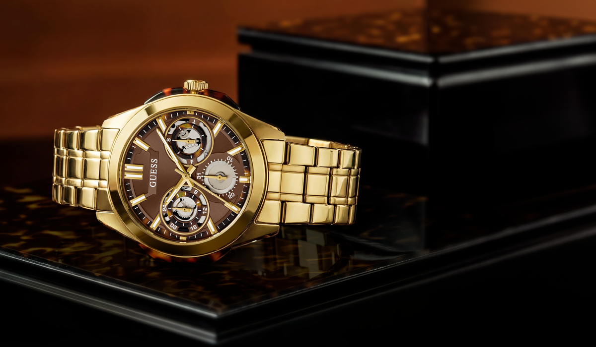 A gold-tone Guess wristwatch with a brown dial and multi-dial face, resting on a glossy black display surface.