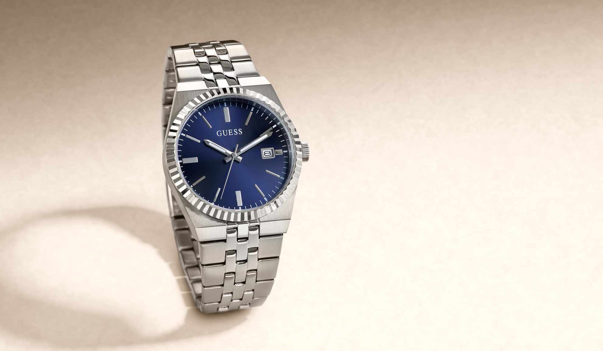 A Guess wristwatch with a silver stainless steel link bracelet and a deep blue dial, shown on a warm beige background.