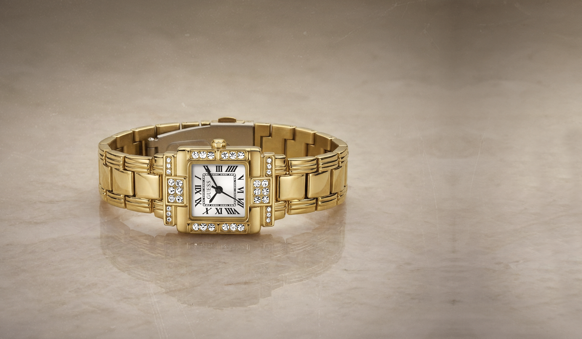 A gold-tone GUESS Tank watch with a diamond-encrusted bezel and a white dial, displayed on a neutral beige marble surface.