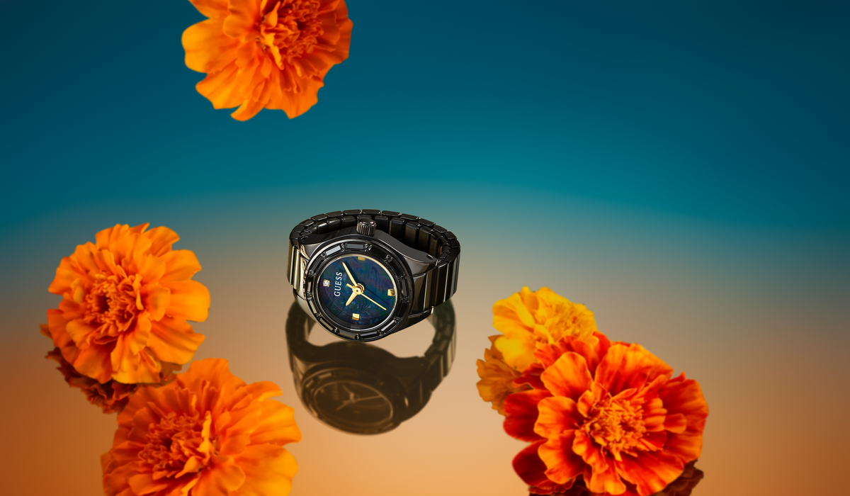 A black analog watch with a blue dial and gold hands sits on a reflective surface, surrounded by bright orange marigold flowers against a blue-to-orange gradient background.