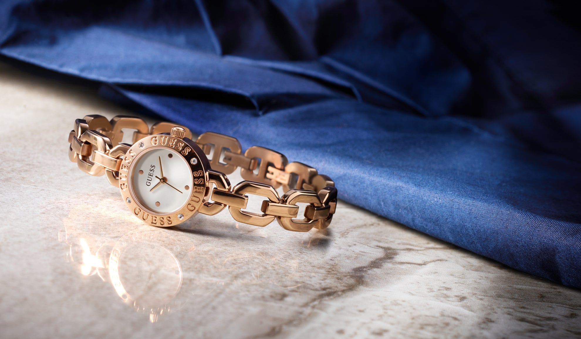 Gold chain-link GUESS watch on a reflective surface with a blue fabric background