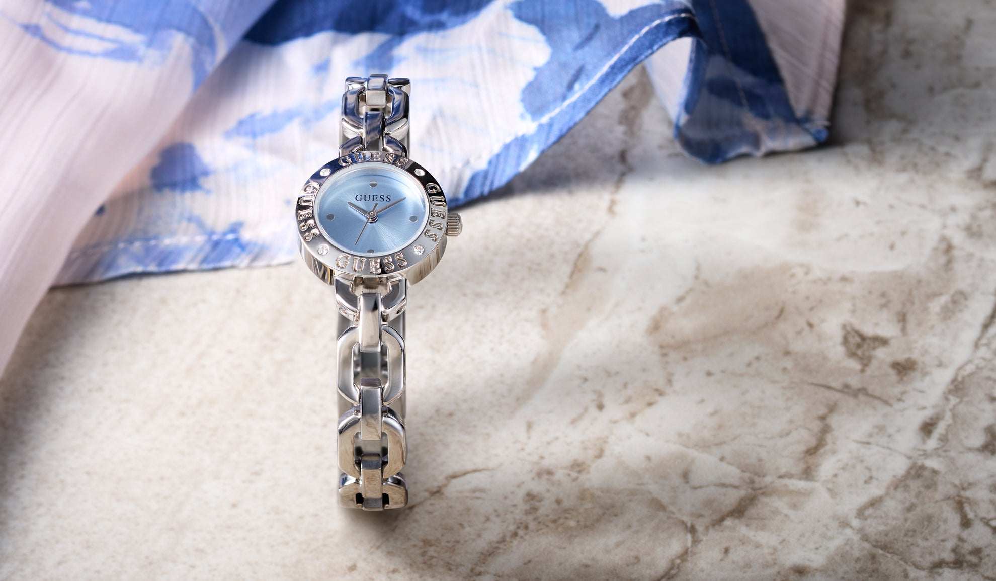 Silver watch with a blue face on a marble surface with a blue and white fabric background