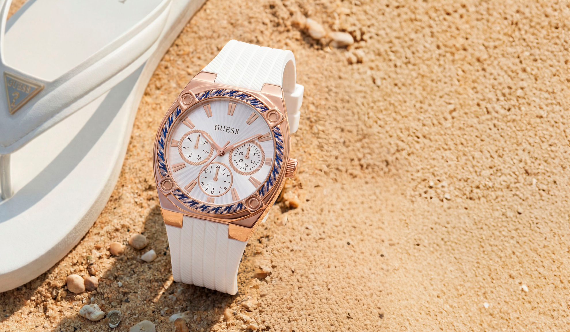 Guess watch with white strap on sand next to white sandals