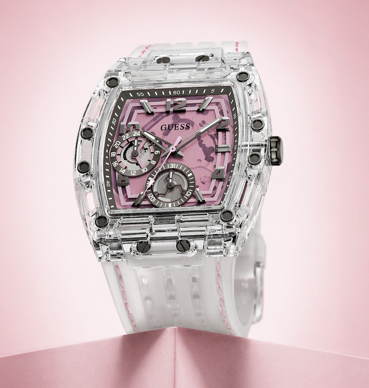 A GUESS watch with a transparent clear case, a pink dial, and a white strap with pink stitching, displayed on a pink background.