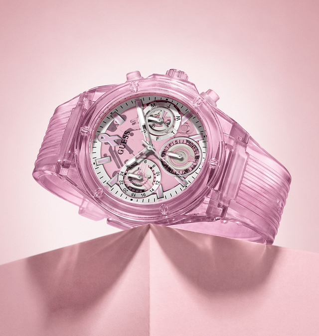 A pink translucent GUESS watch with a matching pink ribbed strap, displayed on a folded pink surface against a soft pink background.