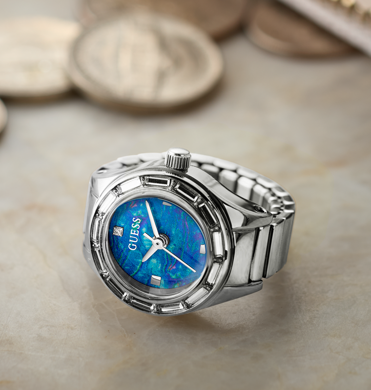A silver-tone stainless steel link watch ring with a blue iridescent dial, resting on a light beige stone surface with blurred coins in the background.