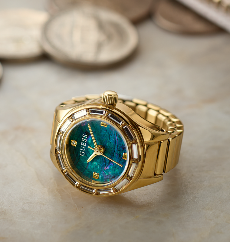 Gold-tone Guess ring watch with a blue-green mother-of-pearl dial and bezel details, resting on a light marble surface with blurred coins in the background.