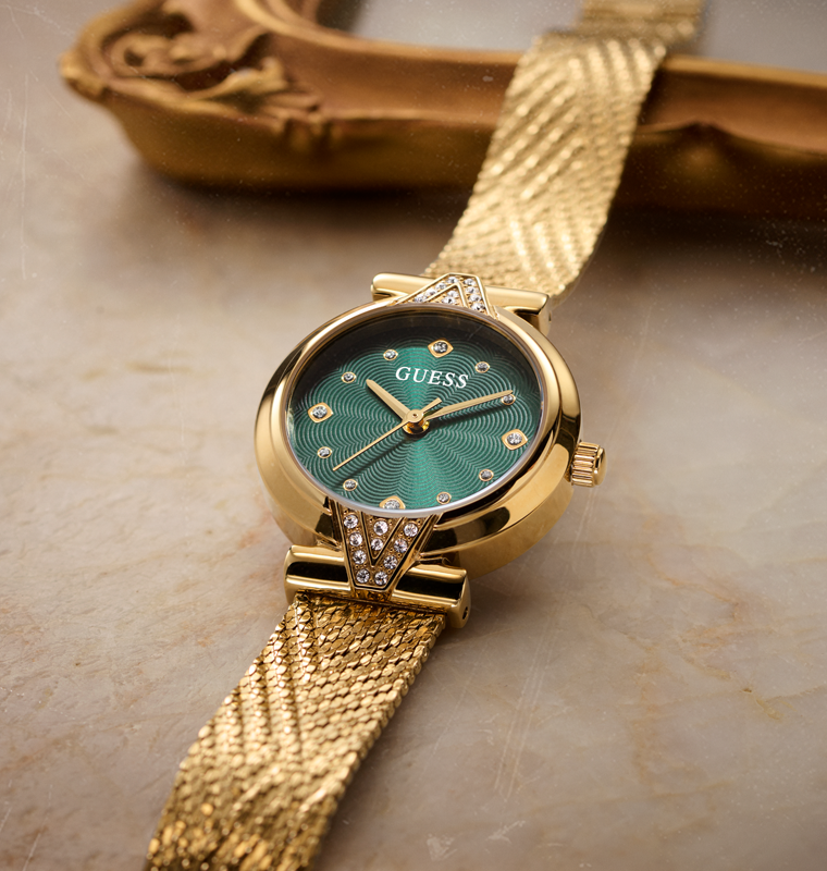 A gold-tone Guess wristwatch with a textured green dial and a matching gold mesh strap, resting on a beige marble surface with a decorative gold object in the background.