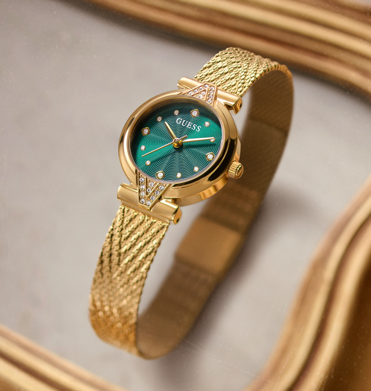 Gold-tone GUESS bracelet watch with a textured mesh band and a green dial, shown on a neutral gray background with a gold reflective border.