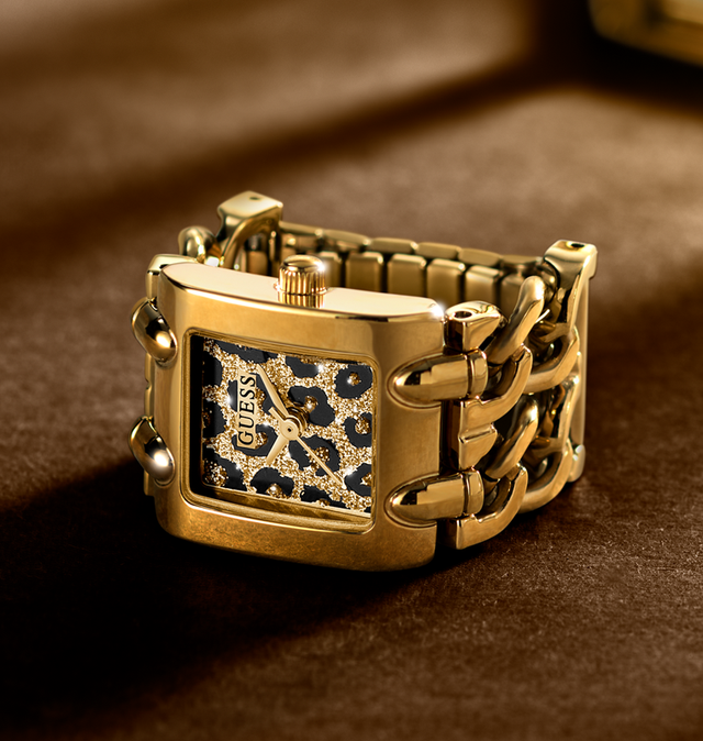 A gold-tone rectangular Guess watch ring with a leopard-print dial and a chunky chain link bracelet, resting on a dark brown textured surface.