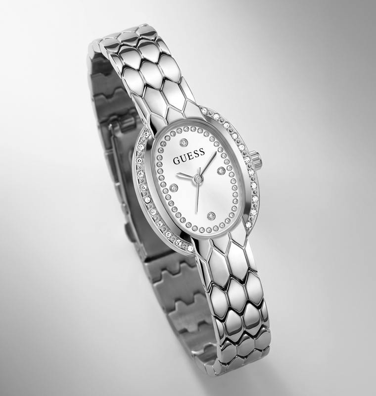 A silver-tone Guess women's bangle watch with a textured honeycomb link bracelet, a silver oval dial, and a bezel set with small clear crystals, shown on a light gray background.