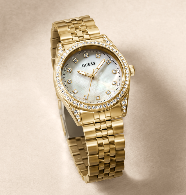 Gold-tone Guess wristwatch with a mother-of-pearl dial, crystal-encrusted bezel, and matching gold-tone link bracelet, photographed on a warm beige background.