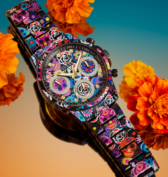 A wristwatch with a black case and a colorful Day of the Dead patterned strap, styled with orange marigold flowers against a blue-to-orange gradient background.