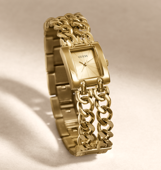 Gold-tone GUESS watch with a rectangular dial and a chunky chain-link bracelet, photographed on a warm beige background with soft shadows.