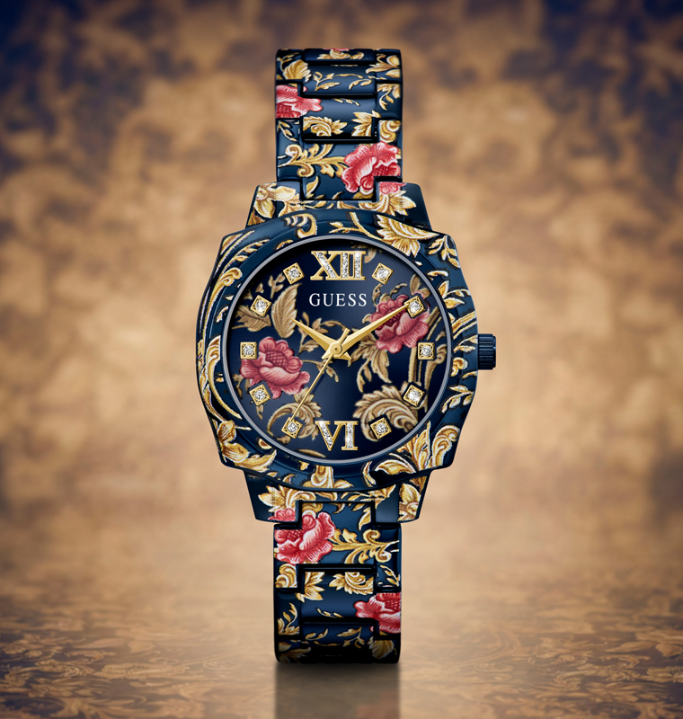 A GUESS women's watch featuring a dark blue dial and a matching patterned bracelet with gold floral motifs and pink rose accents, set against a textured brown background.