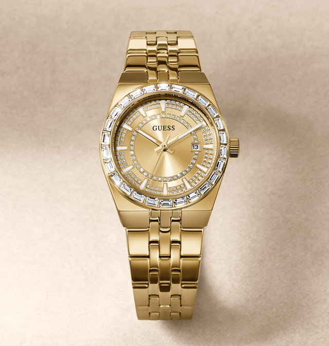 A gold-tone Guess wristwatch with a bezel set with clear crystals and a matching crystal-accented dial, shown on a neutral beige background.