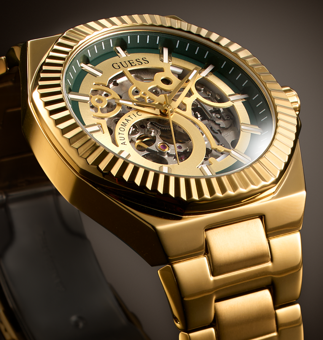 Gold-tone GUESS automatic skeleton watch with a green dial and gold-tone metal bracelet, shown in a close-up studio shot.