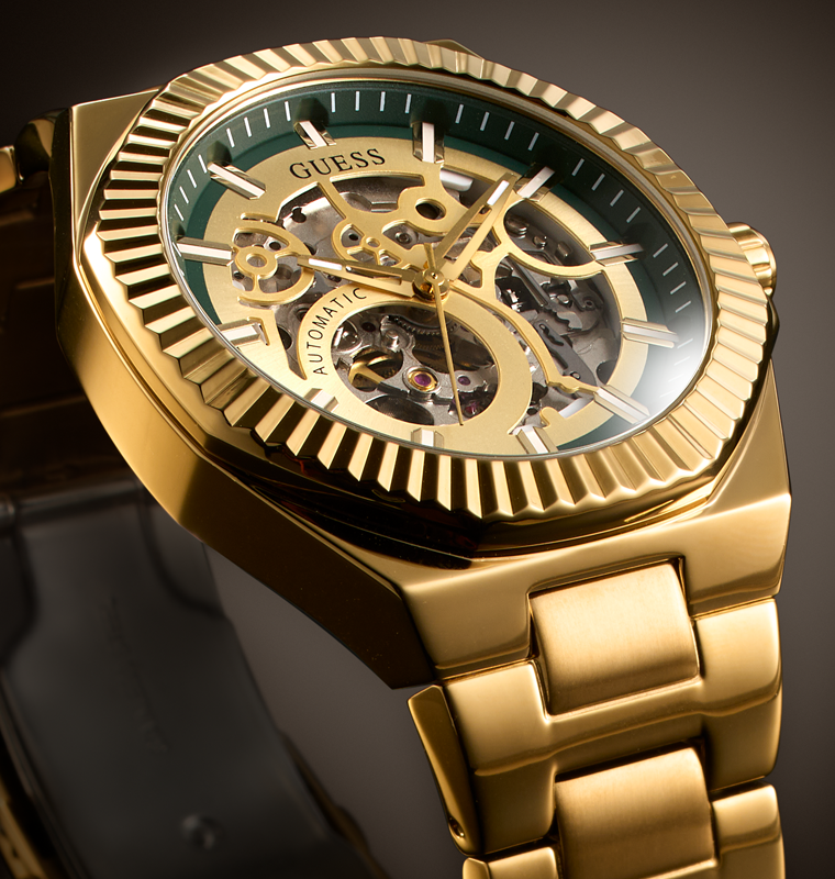 Gold-tone GUESS automatic skeleton watch with a green dial and gold-tone metal bracelet, shown in a close-up studio shot.