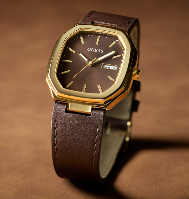 A GUESS wristwatch with an octagonal gold-tone case, a dark brown dial, and a brown leather strap, shown on a warm brown textured background.