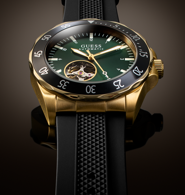 A luxury wristwatch by Guess with a gold-tone case, black bezel, dark green dial, and a black textured strap, shown in a close-up studio shot.