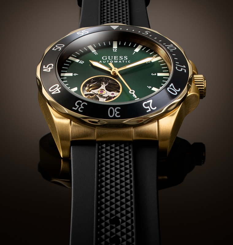 A luxury wristwatch by Guess with a gold-tone case, black bezel, dark green dial, and a black textured strap, shown in a close-up studio shot.