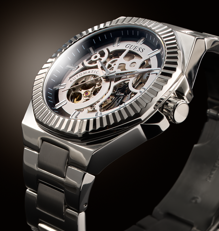 A close-up shot of a GUESS automatic skeleton watch with a stainless steel case, fluted bezel, and open-worked dial showing the inner mechanical movement, set against a dark background.