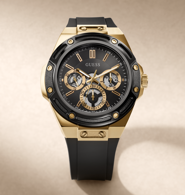 A luxury wristwatch by Guess with a gold-tone case, black bezel and dial, and a black rubber strap, shown on a neutral beige background.