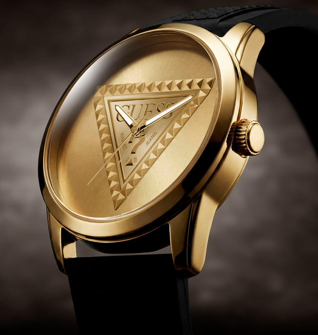 A close-up shot of a Guess men's watch featuring a gold-tone case, a gold dial with an embossed triangle logo, and a black strap, set against a dark textured background.