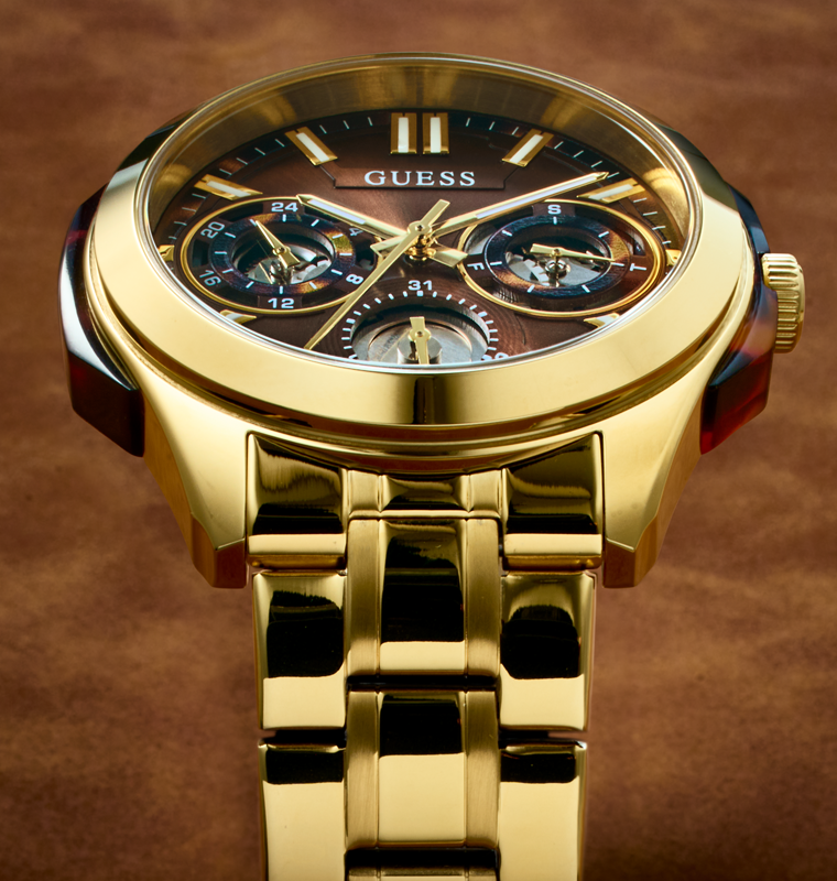 Gold-tone Guess wristwatch with a matching metal link bracelet, featuring a dark brown dial with chronograph subdials and gold-tone hands and markers.