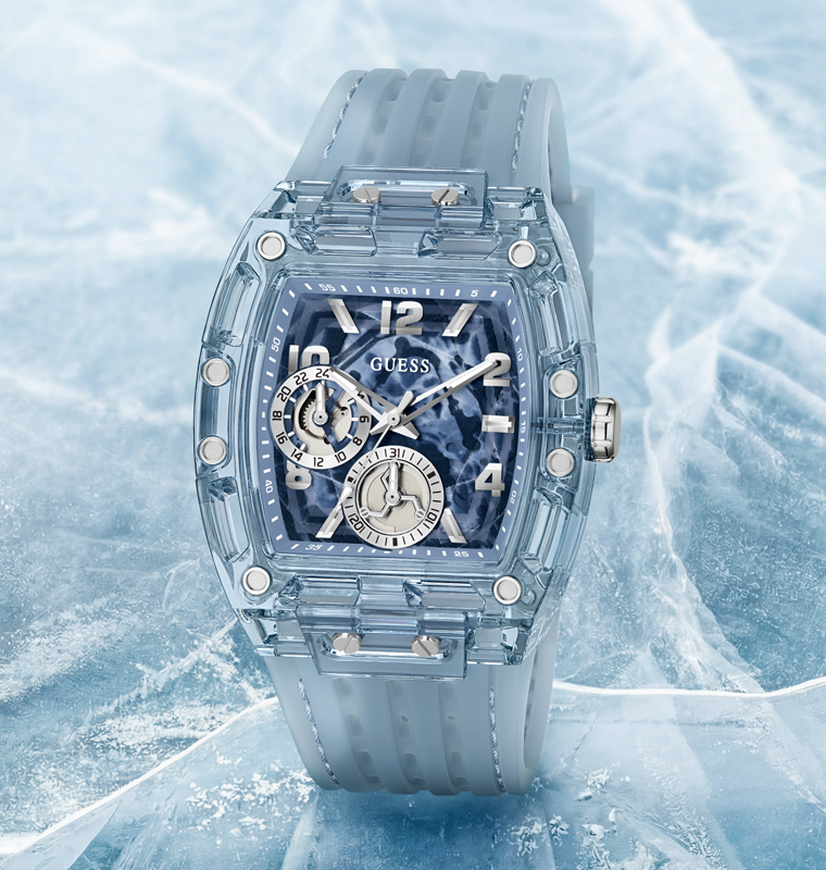 A Guess watch with a translucent light blue case and matching strap, featuring a dark blue marbled dial with silver-tone details, set against a textured icy background.